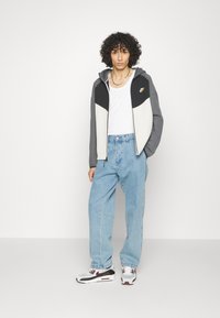 Felpa con zip da uomo in grigio, nero e crema con un logo Nike ben visibile, abbinata a jeans baggy azzurri e sneakers bianche.