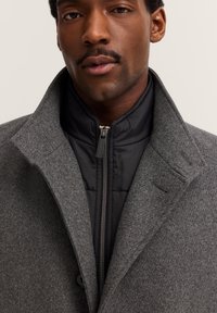 Grau wollener Übermantel mit Reverskragen, über einer schwarzen gepolsterten Jacke mit Reißverschluss und glatter Textur sowie glänzenden Hardware-Elementen.