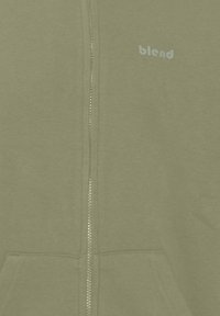 Felpa olive green con zip realizzata in tessuto morbido, dotata di due tasche anteriori e di un sottile logo "blend" in azzurro chiaro sul petto.