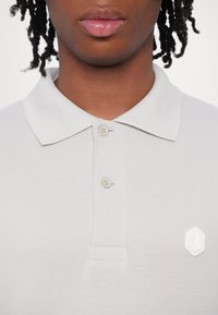 Světle šedé polo tričko s texturovanou tkaninou, klasickým límcem, dvěma knoflíky a malým bílým geometrickým logem na hrudi.