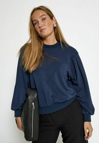 Navy blue pullover with a V-shaped design, long puff sleeves, and a ribbed neckline, paired with a black leather handbag featuring a zipper.