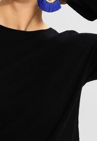 Musta neulepaita, jossa on pyöreä kaula-aukko, yhdistetty suuriin sinisiin tupsukorvakoruihin, joissa on pyöreä puinen koru.