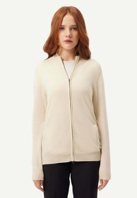 Beige zip-up kofta gjord av mjukt tyg, med lång ärm, ribbade muddade ärmar, två sidofickor och en hög krage.