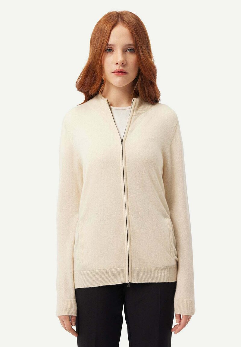 Beige zip-up kofta gjord av mjukt tyg, med lång ärm, ribbade muddade ärmar, två sidofickor och en hög krage.