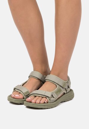 Füße, die hellgraue Sandalen mit verstellbarem Riemen und dicker Sohle auf einem hellgrauen Hintergrund tragen.