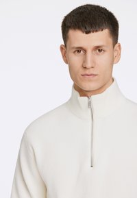 Hvid ribbet pullover med høj krave og halv-zip lukning. Har et struktureret stof og minimalistisk design.
