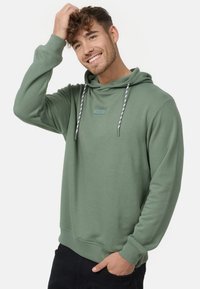 Grüne Kapuzenjacke aus weichem Material mit Kordelzug und einem kleinen Aufnäher auf der Vorderseite. Verfügt über gerippte Bündchen und Saum, kombiniert mit schwarzen Hosen.
