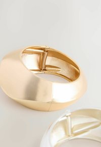 Guld bangle armbånd med et glat, buet design, metallisk finish og en synlig hængselmekanisme til åbning og lukning.