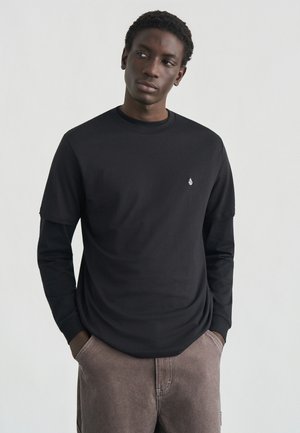 T-shirt noir à manches longues avec col rond côtelé et petit logo blanc sur la poitrine, associé à un pantalon marron texturé.