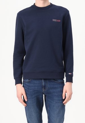 Person trägt marineblauen Langarm-Sweatshirt mit Rundhalsausschnitt und kleinem Logo auf Brust und Handgelenk, kombiniert mit blauen Denim-Jeans, steht vor weißem Hintergrund.
