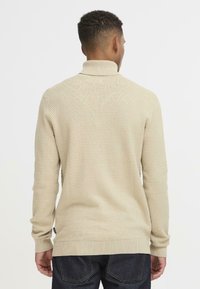 Beige textúrájú pulóver magas gallérral, bordázott mandzsettákkal és aljjal, diszkrét mintával. Sötét farmernadrággal viselve.