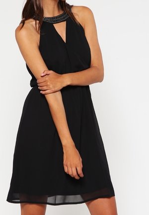 Donna che indossa un vestito nero senza maniche con colletto decorato con perline, che incrocia un braccio sull'altro davanti a uno sfondo bianco uniforme.