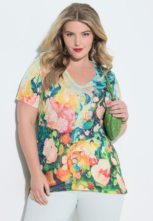 Frau mit blondem Haar trägt buntes, geblümtes Hemd, weiße Hose, roten Lippenstift, Ohrringe und hält eine kleine grüne Handtasche vor weißem Hintergrund.