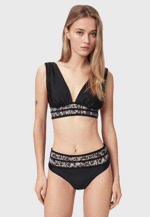 Kvinde med langt blondt hår har en sort bikini med dyreprint-bånd på og står foran en ensfarvet lysegrå baggrund.
