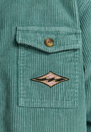 Teal corduroy-paita, jossa on rintatasku napilla, jota koristaa timantin muotoinen brodeerattu logo vaaleanpunaisena ja mustana.