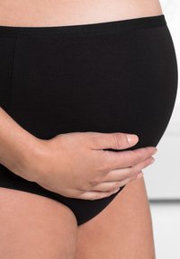 Slip di cotone nero con una texture liscia, dotati di un elastico in vita. Una mano poggia su una pancia rotonda e sporgente.