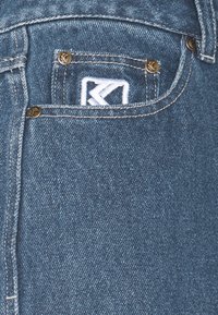 Denimblå jeans med en sydd bakficka med en vit logo, guldfärgade nitar och kontrasterande stygn längs sömmarna.