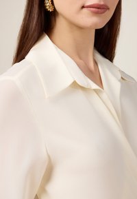 Cremfarbene Bluse mit einem Kragenoberteil und weicher, glatter Textur. Verfügt über ein Wickeldesign und subtilen Glanz. Goldene Creolen unterstreichen den Look.
