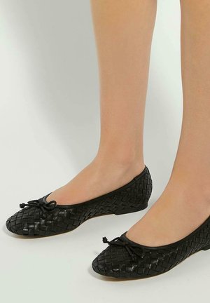 Voeten met zwarte geweven leren ballerina's met kleine strikaccenten op een effen lichte achtergrond.