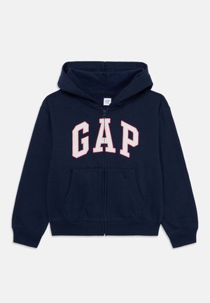 Granatowa bluza z kapturem zapinana na zamek, z przednimi kieszeniami i dużymi białymi literami "GAP" obramowanymi na różowo na piersi.