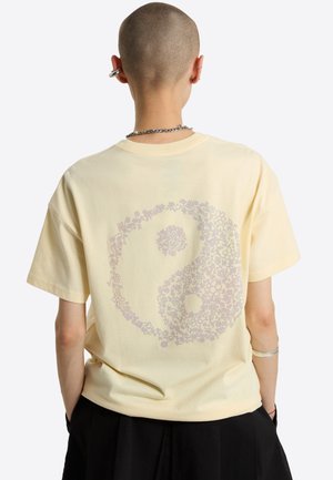 Persona con la cabeza rapada que lleva una camiseta amarilla pálida con un diseño floral de yin-yang en la espalda, pantalones negros y joyería plateada.