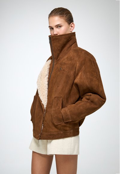 Jachetă din suede de culoare maro, cu guler înalt, având o căptușeală moale din blană și două buzunare frontale. Închidere cu fermoar și design confortabil.
