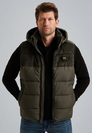 Homme portant un gilet rembourré vert olive avec une capuche et un t-shirt �à manches longues noir, se tenant les mains dans les poches devant un fond uni.