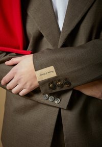 To kryssede armer iført brune dressjakker med knappedetaljer, det ene ermet viser "House of Sunny"-etikett, rød bakgrunnsstoff.