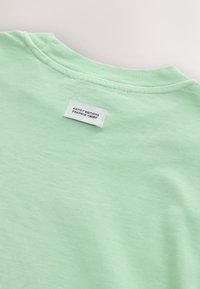 Ljusgrön bomullst-shirt med rund halsringning, med en liten vit etikett med svart text sydd vid nackens baksida.