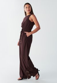 Ruda rankovių neturinti kombinezonas su elastinga juosmens juosta ir šoniniais kišenėmis; priderinami raišteliai prie kulkšnių su auksinėmis detalėmis.