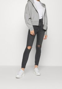Grå zip-up hoodie, vit ribbad tröja, svarta skinny jeans med knäskärningar och vita plattformssneakers. Avslappnad outfit med moderna inslag.