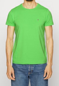 Vihreä puuvillainen T-paita, jossa on pyöreä kaula-aukko, lyhyet hihat ja pieni brodeerattu logo rinnassa. Yhdistetty sinisiin farkkuihin.