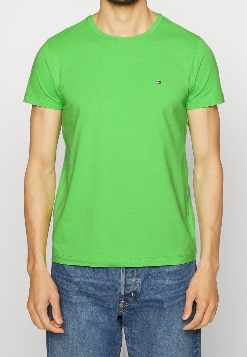 Vihreä puuvillainen T-paita, jossa on pyöreä kaula-aukko, lyhyet hihat ja pieni brodeerattu logo rinnassa. Yhdistetty sinisiin farkkuihin.