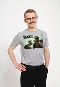 Šedé bavlněné tričko s potiskem portrétu z "Útěku z New Yorku", zelenými akcenty a černým textem. Model má na sobě černé kalhoty.