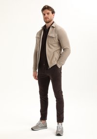 Hombre de pie con chaqueta beige con cremallera, pantalones marrón oscuro, zapatillas grises y camiseta negra, con las manos en los bolsillos, contra un fondo blanco.