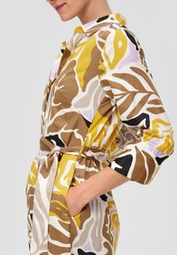 Robe chemise multicolore avec un motif abstrait audacieux en jaune, marron, noir et lavande ; elle possède une taille ceinturée et des poches latérales.