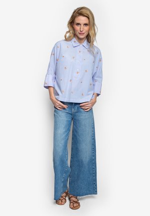 Frau in einer locker sitzenden blauen Bluse mit kleinen orangefarbenen Blumen, weit geschnittenen blauen Jeans und braunen Riemchensandalen, die vor einem weißen Hintergrund steht.