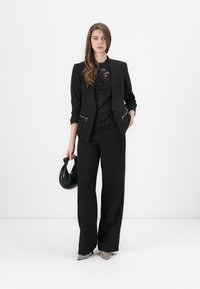 Blazer nero su misura con tasche con zip, sovrapposto a un top in pizzo, abbinato a pantaloni neri a gamba larga e in mano a una borsa nera.