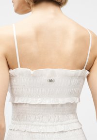 Άνω μέρος της πλάτης γυναίκας που φοράει ένα λευκό ρούχο με σφηκοφωλιά, λεπτές τιράντες και μικρό μεταλλικό λογότυπο "MK" στο πάνω μέρος της πλάτης.