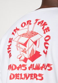 Bílá mikina s červeným grafickým potiskem krabičky na nudle s hůlkami a textem "STRAVUJTE SE NEBO S SEBOU" a "ADIDAS VŽDY DODÁVÁ."