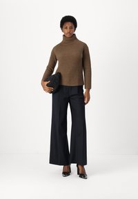 Gestrickter brauner Rollkragenpullover, weite schwarze Hose und schwarze High-Heels. Die Modells hält eine strukturierte schwarze Clutch.