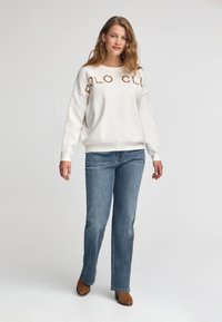 Frau trägt einen weißen Pullover mit dem Text "POLO CLUB", blaue Jeans und braune Stiefeletten, steht vor einem schlichten hellen Hintergrund.