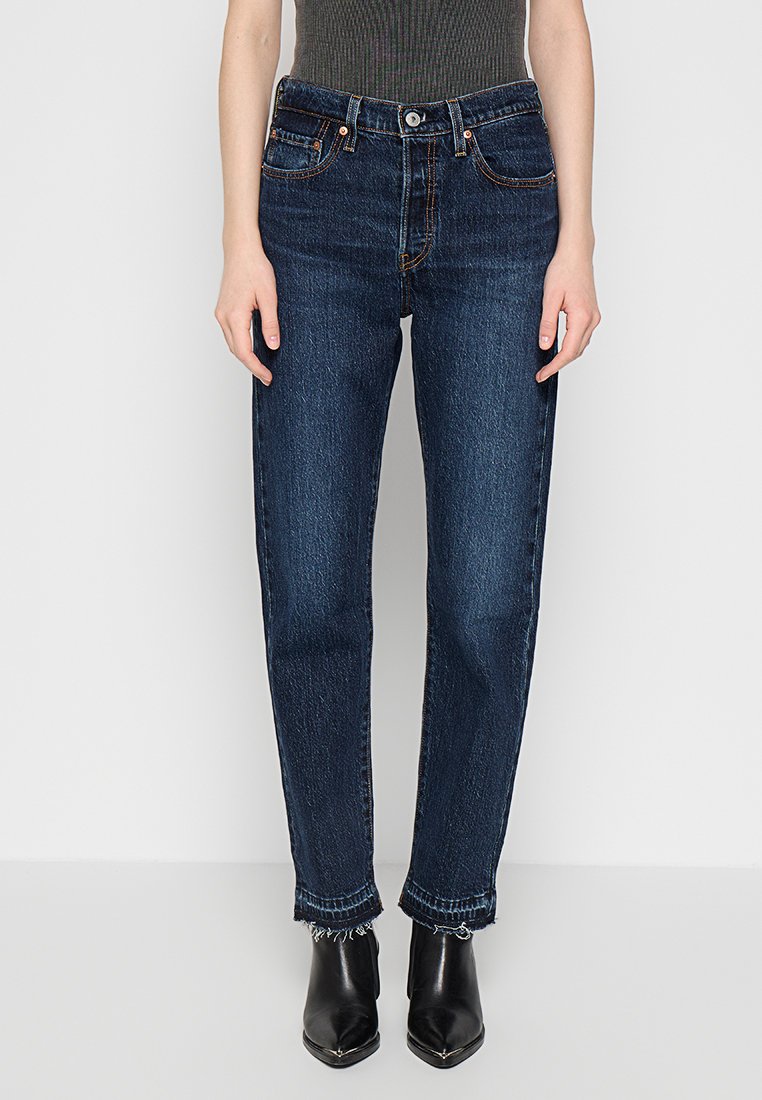 Jean en denim bleu foncé avec une coupe droite et ample. Caractéristiques : ourlet effiloché, design à cinq poches et coutures contrastantes. Associé avec des bottines noires.