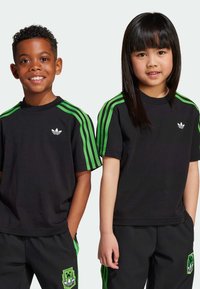 Svart T-shirt med gröna randiga detaljer, med en liten vit logotyp på bröstet. Matchas med svarta byxor och gröna detaljer på sidorna.