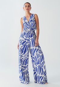 Plavi i bijeli kombinezon uzorka s omotnim dizajnom, širokim nogavicama i vezicom u struku. Bez rukava s dubokim V-izrezom. Lagani materijal.