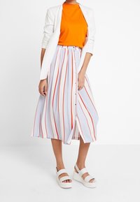 Oranžové tričko, bílý blejzr, světle modrá sukně s červenými svislými pruhy a knoflíkovým detailem vpředu, doplněné robustními bílými sandály.