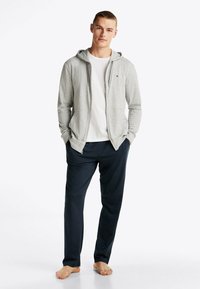 Sudadera gris con cremallera ycordones sobre una camiseta blanca, combinada con pantalones de chándal azul oscuro. Tejido suave y corte relajado.