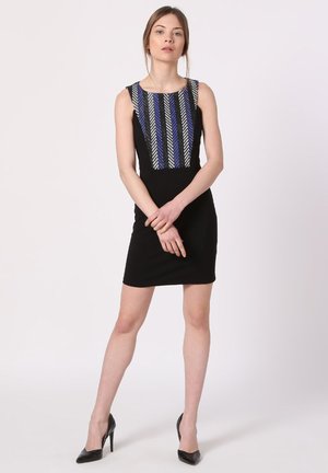 Mujer de pie con las manos cruzadas, vistiendo un vestido negro sin mangas con un patrón de rayas verticales en el pecho y tacones altos negros.