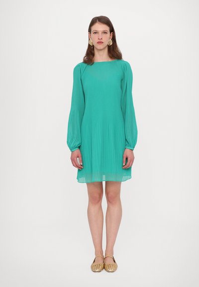 Een turquoise geplooide jurk met lange, geplooide mouwen, een vierkante halslijn en een vloeiende silhouet, gecombineerd met gevlochten gouden flats.