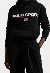 Schwarzer Kapuzenpullover mit "POLO SPORT" in weißer und roter Schrift, ausgestattet mit einer kleinen amerikanischen Flagge, aus weichem Stoff, mit einer Fronttasche.